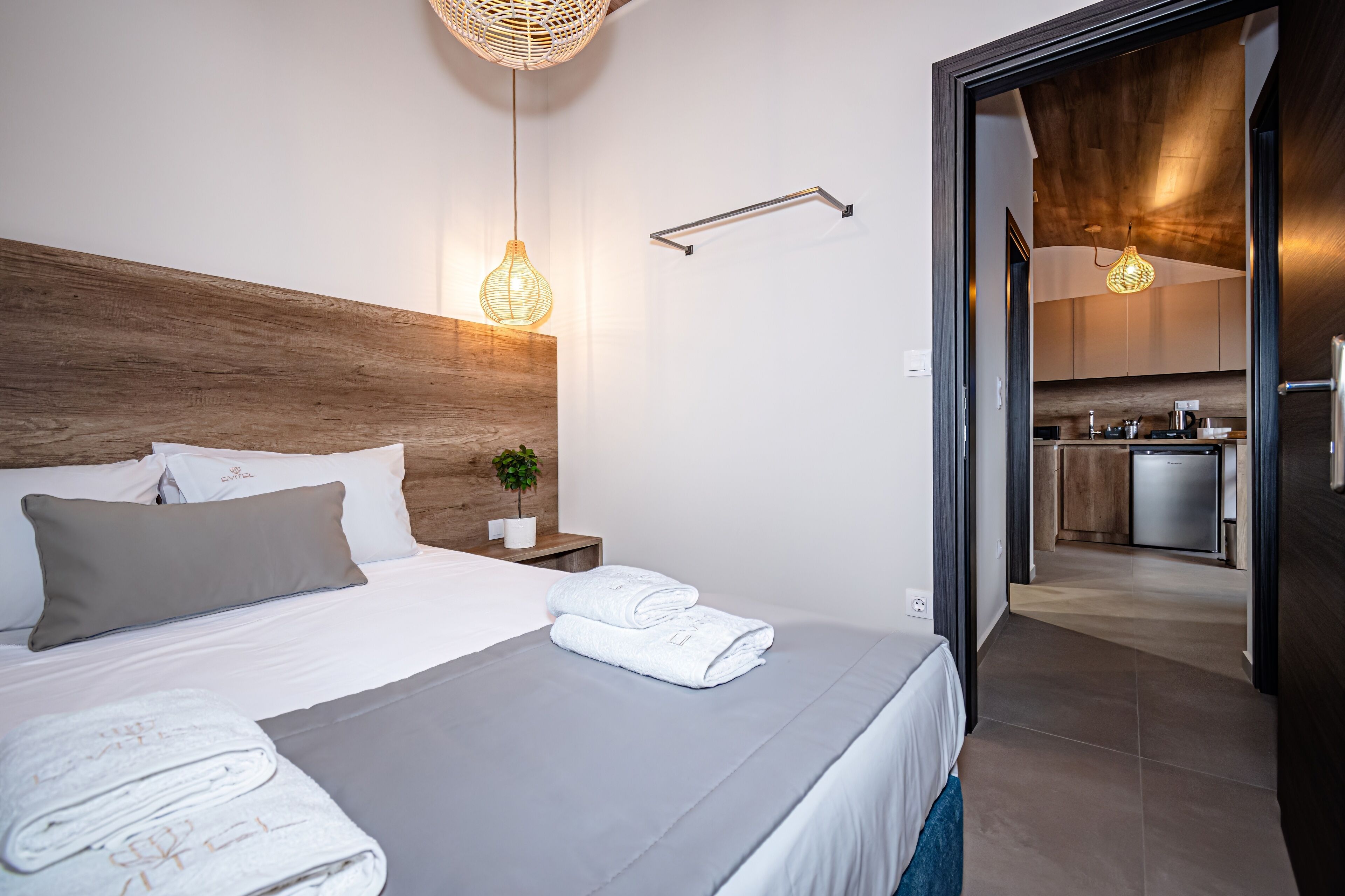 A modern hotel room with a wooden headboard, a bed with white linens, a grey blanket, and a neatly made bed with towels. There is a small bedside table with a plant and a wall-mounted towel rack.