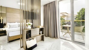 A modern hotel room with a bed, a bedside table with a lamp, a large window with curtains, and a balcony with outdoor furniture. - Evitel Luxury Living (Kassandra)