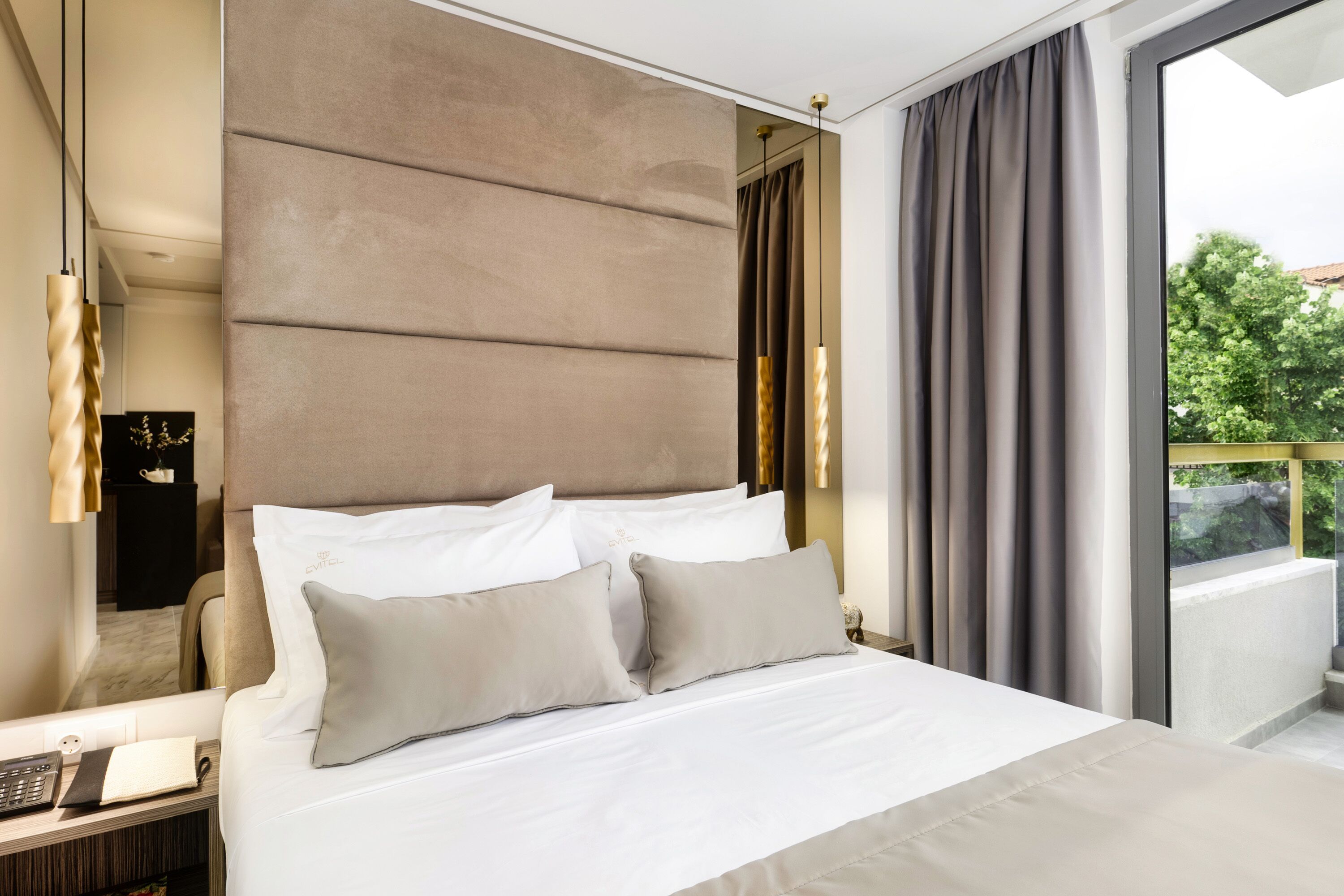 A modern hotel room with a large bed, a bedside table with a lamp, and a view of the outdoors through a large window.