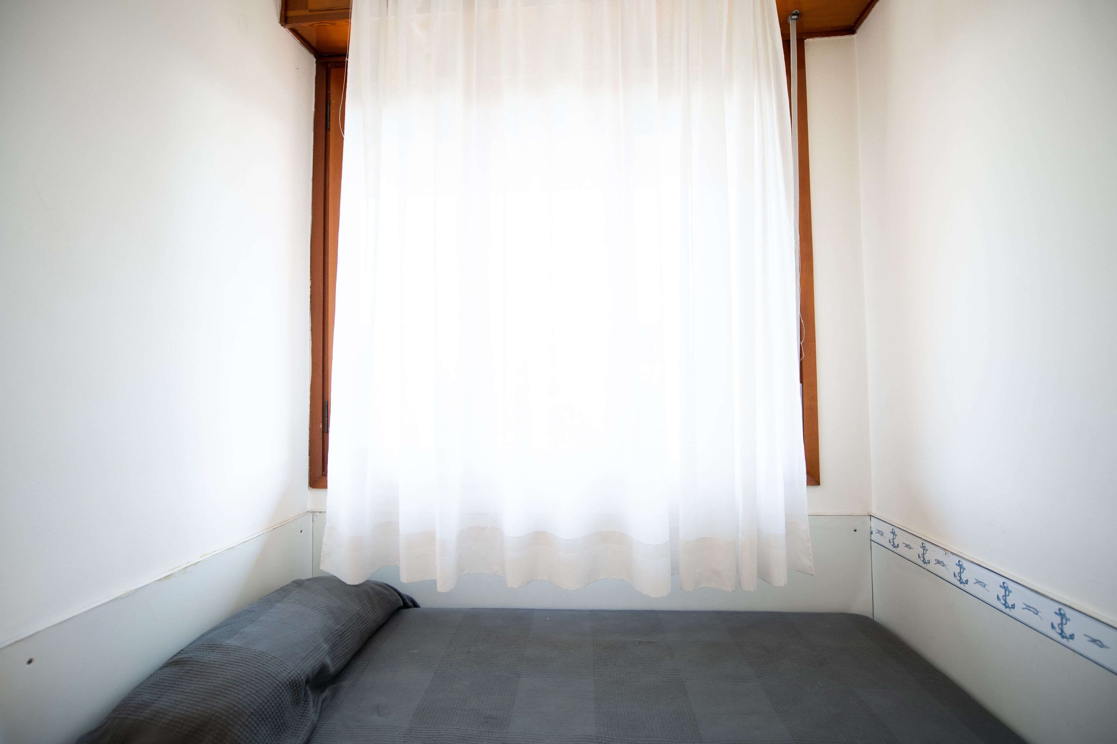 Een kleine kamer met een enkel bed, een effen witte muur, een houten plafond en een raam met dunne gordijnen.