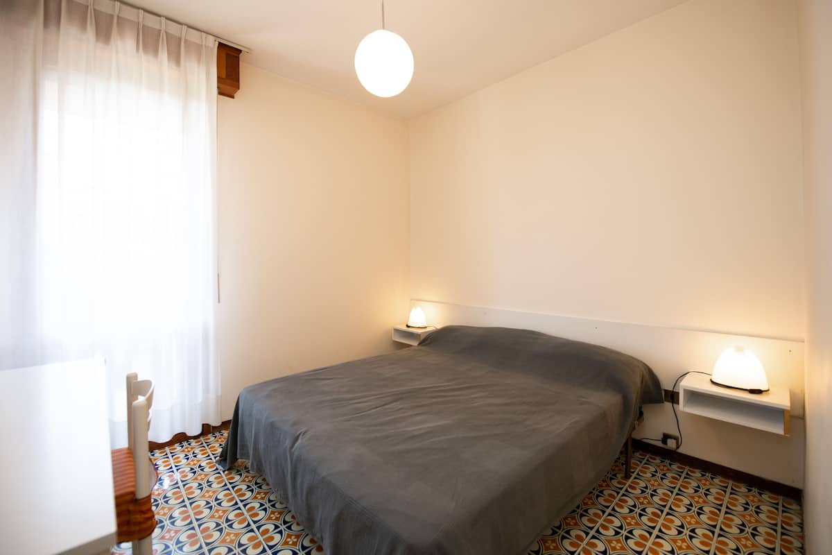 Een slaapkamer met een bed, nachtlampjes, een raam met gordijnen en vloertegels met een patroon.