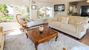 Een ruime woonkamer met een groot raam, een witte bank, een houten salontafel en een rieten fauteuil.