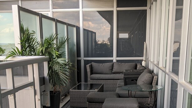 Un espacio de estar al aire libre cubierto con piso de madera oscura, mobiliario de mimbre gris y una mesa de vidrio.