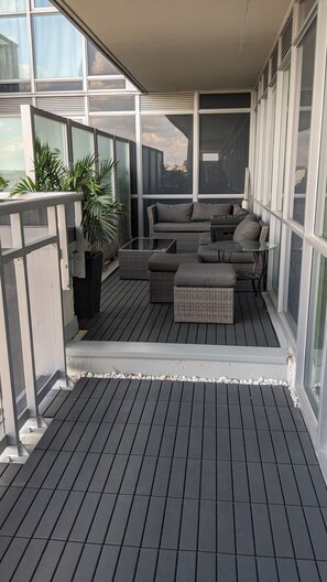 A covered outdoor seating area with a dark wooden floor, grey wicker furniture, and a glass table. - Luxury Condo- GO station, GYM, Pool, BBQs (Burlington)