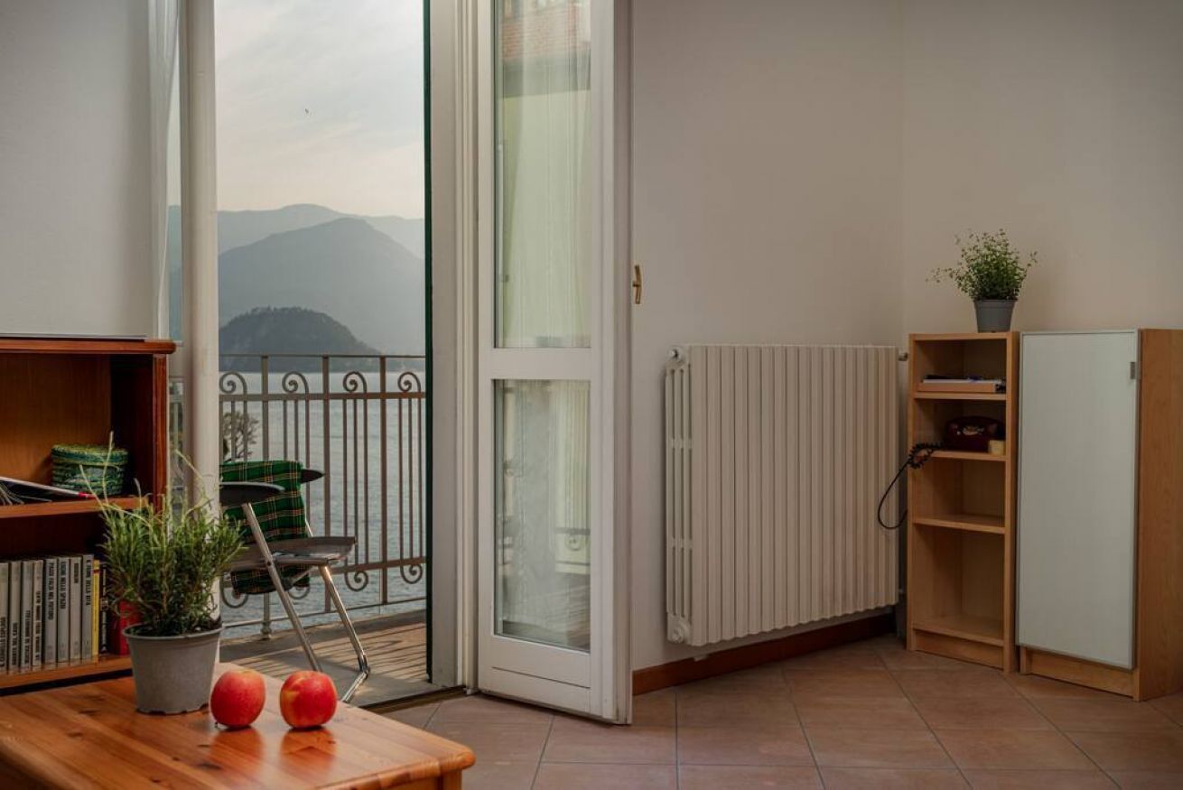A balcony with a view of mountains, a radiator, a wooden table with apples and a plant, and a bookshelf.