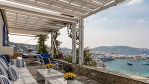 Une terrasse dotée d’une pergola blanche, de canapés à rayures bleues et blanches, et offrant une vue sur une ville côtière et un port.