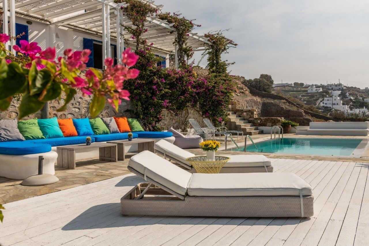 A rooftop pool area with lounge chairs, a sofa with colorful cushions, and a view of a hillside with white buildings.
