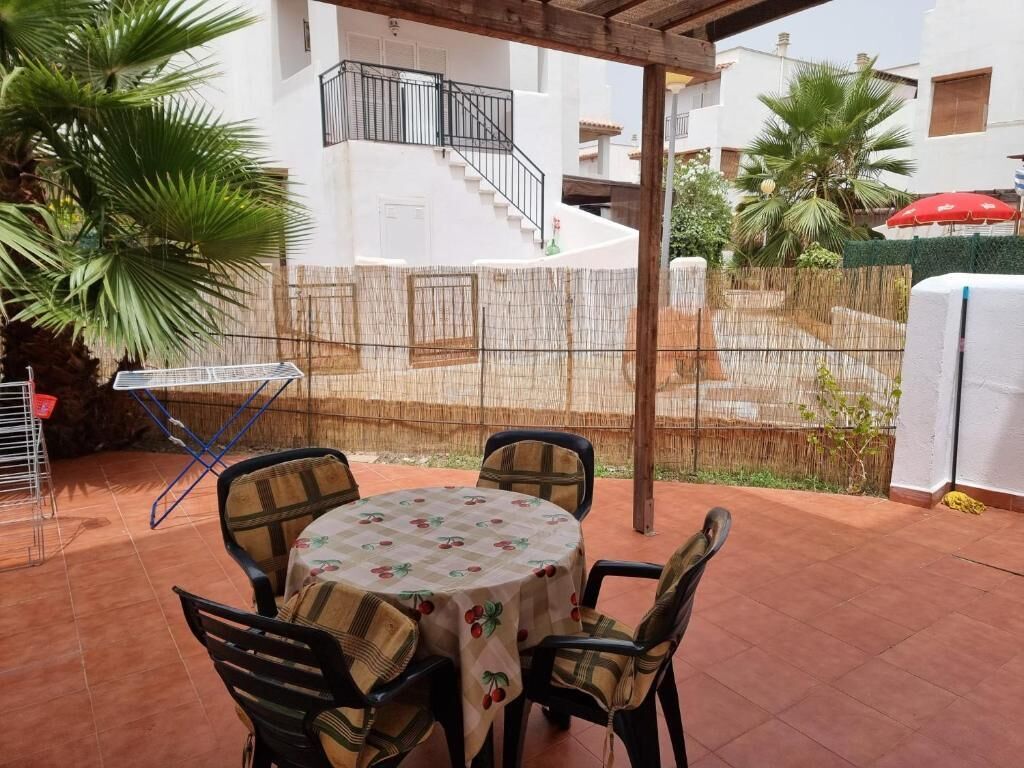 Une terrasse extérieure avec une table ronde et des chaises, un espace salon couvert et une vue sur un bâtiment blanc doté d’un escalier.