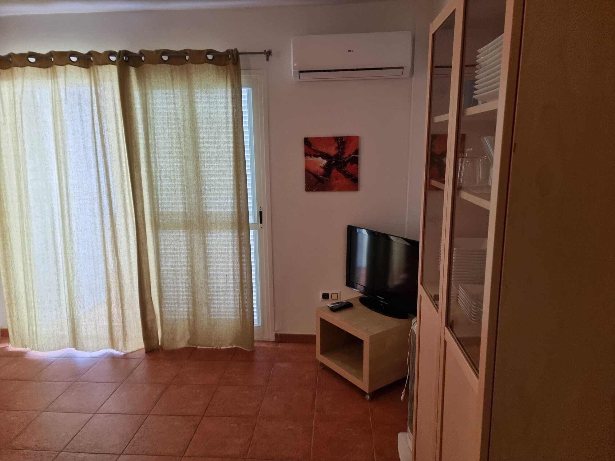 Una sala de estar con televisor sobre un mueble bajo, una ventana con cortinas y un equipo de aire acondicionado en la pared.