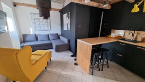 En kompakt opholdsstue med en sofa, en gul stol, et tekøkken med vask og barstole, samt et sort skab.