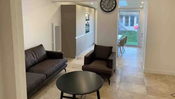 Uma sala de estar moderna com sofá, poltrona e mesa de centro. Há um relógio na parede e uma porta dá para uma varanda.
