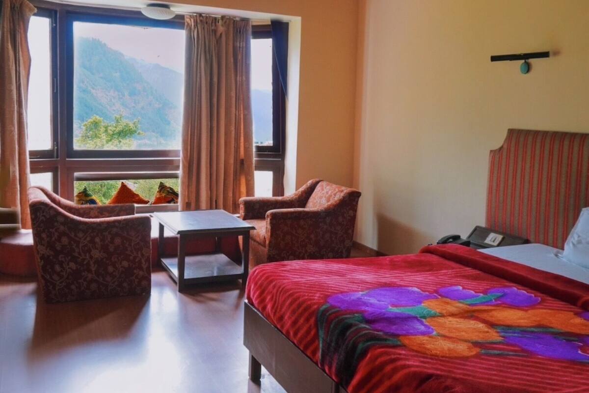A room with a bed, a small table, two chairs, a window with curtains, and a view of mountains.