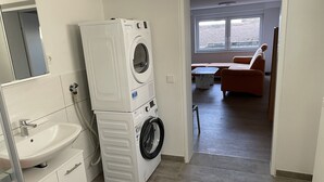 A laundry room with stacked washers and dryers, a sink, and a view into a living area with a sofa and a window. - Vacation apartment in the countryside (Lebach Landsweiler)
