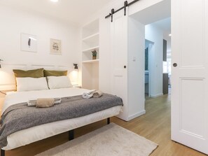 Un dormitorio ordenado con cama, mesitas de noche, estante en la pared y puerta que da a otra habitación.