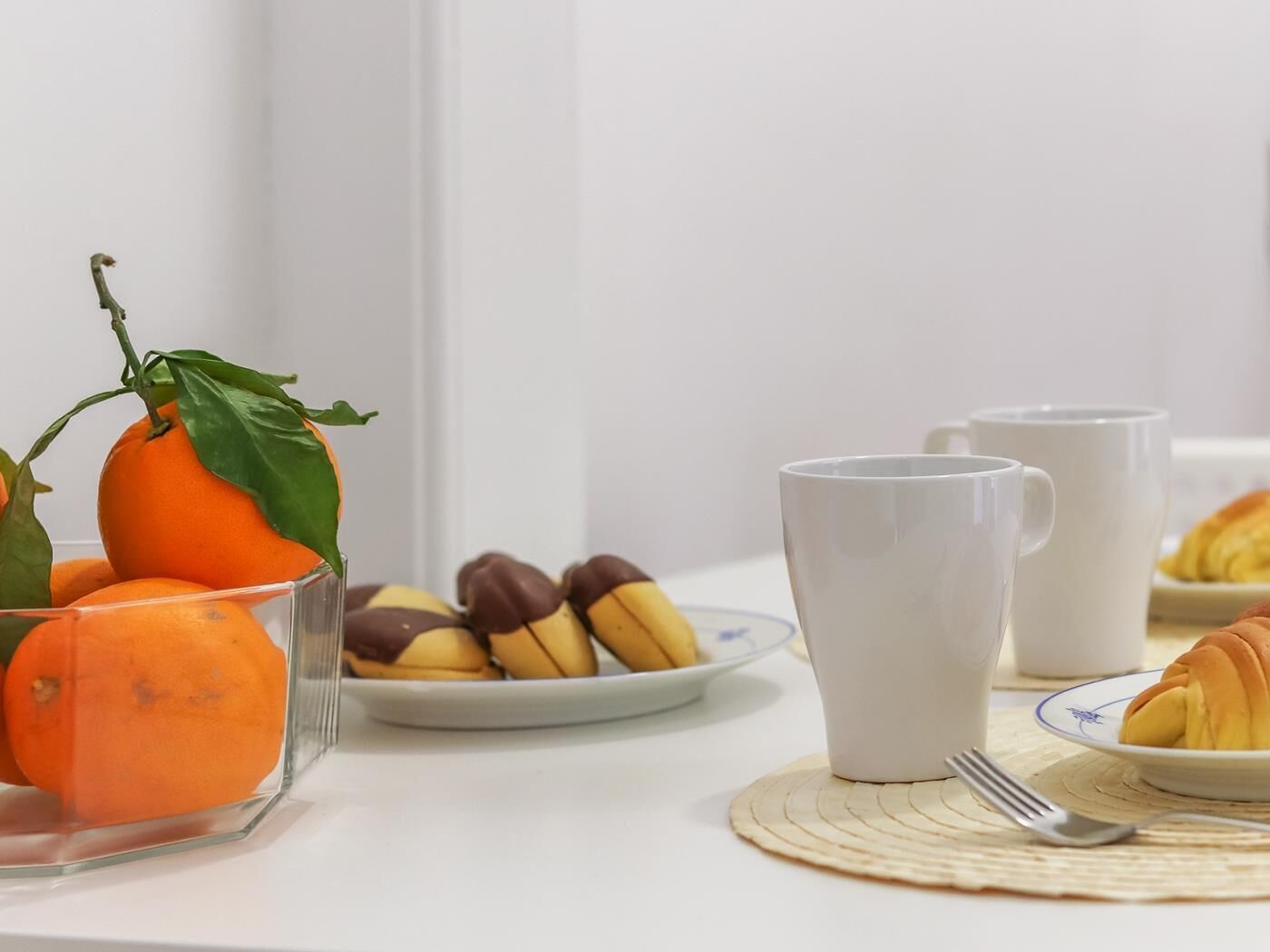 Een ontbijttafel met een glas sinaasappelsap, een bord bananen en hazelnoten, twee witte mokken, een croissant en een vork op een onderzetter.