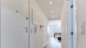En modern hall med vita väggar, trägolv och infällda taklampor. På vänster sida finns två dörrar, varav den ena har ett handtag och den andra ett lås. En liten hylla med en spegel hänger på väggen, och en telefon är monterad på väggen.