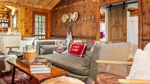 A cozy lodge interior with a wooden interior, a sofa with 'Lola's Lodge' pillows, a dining area with a wooden table, and a kitchen visible through a window. - Chic, updated log cabin nestled in the pine trees of Sprucewold! (Boothbay Harbor)