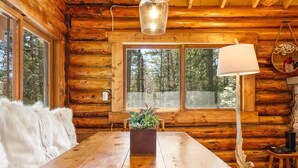 A wooden cabin interior with a dining table, a potted plant, and a window with a view of the forest. - Chic, updated log cabin nestled in the pine trees of Sprucewold! (Boothbay Harbor)