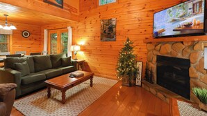 L’intérieur chaleureux d’un chalet en bois, avec une cheminée, un téléviseur, un canapé vert et un sapin de Noël.