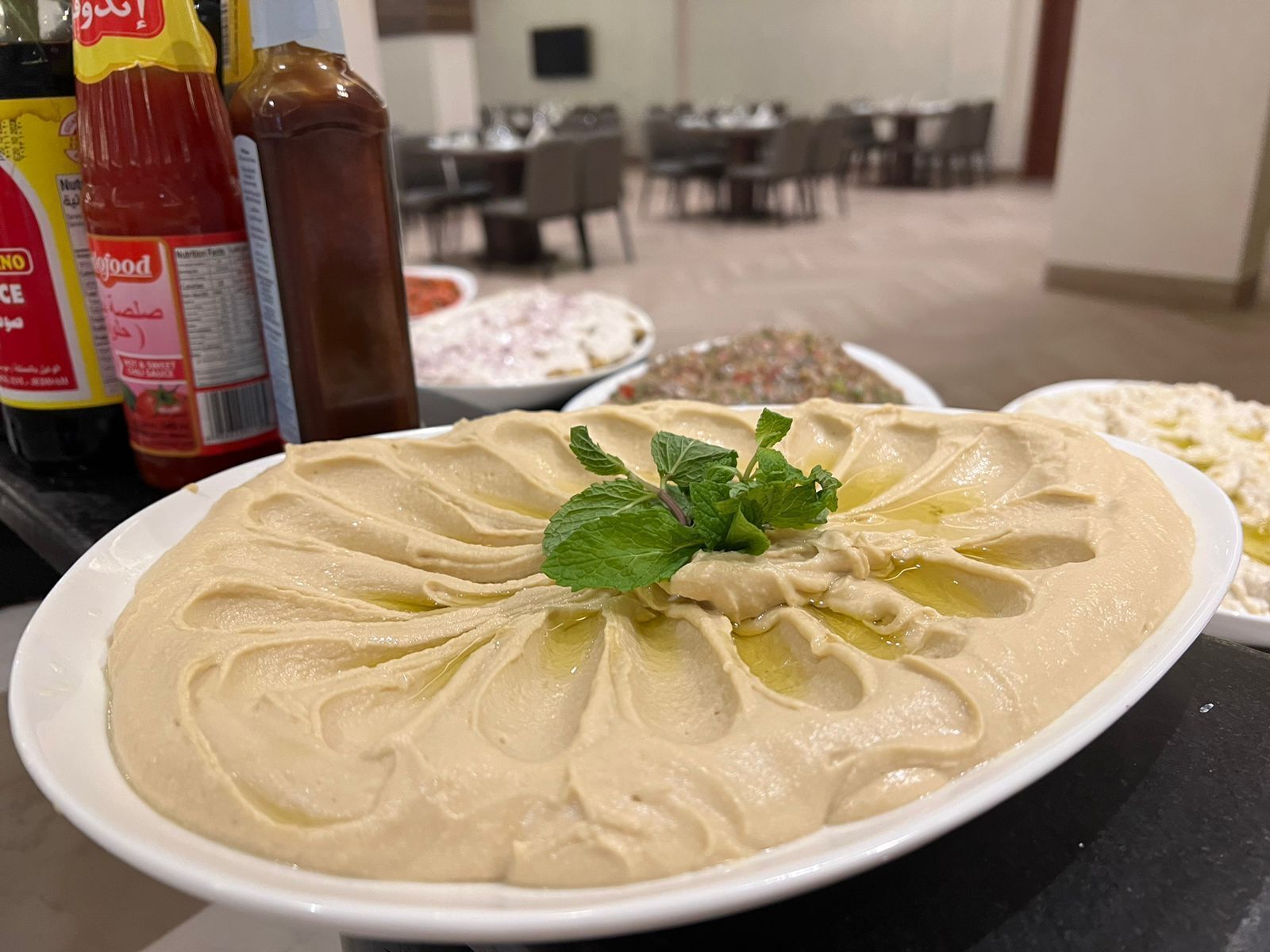 Ein Teller Hummus, garniert mit Minzblättern, auf einem Tisch. Im Hintergrund sind verschiedene Gewürze und Beilagen zu sehen.