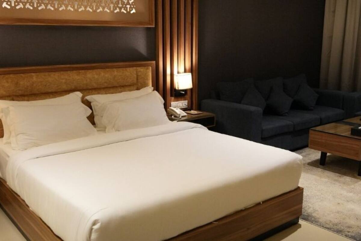 Habitación de hotel con una cama grande, un sofá oscuro, una mesa de centro de madera y una mesita de noche con lámpara.