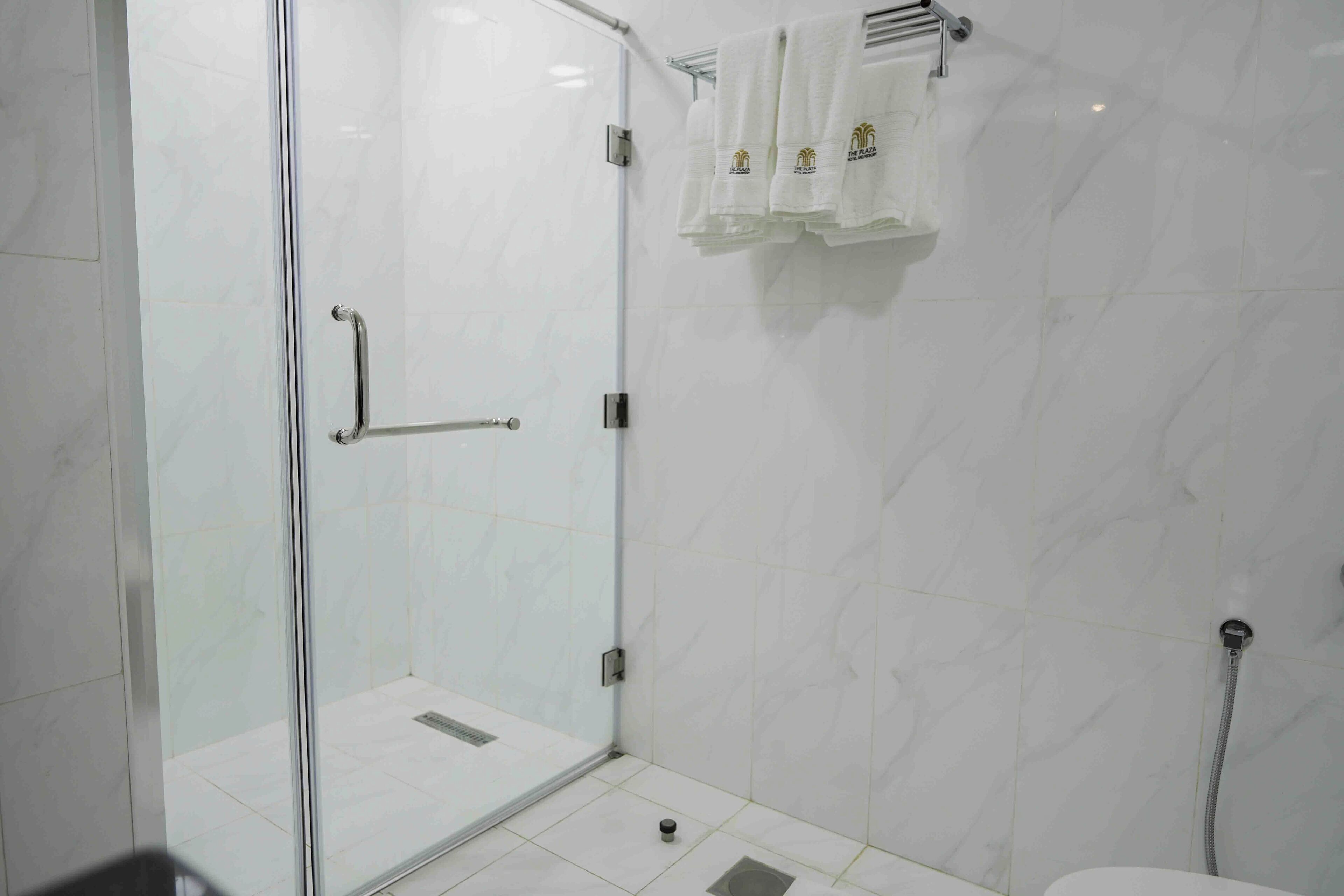 Ducha con mampara de vidrio, barra de apoyo, paredes de azulejos blancos y un perchero con toallas.