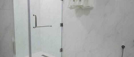Ducha con mampara de vidrio, barra de apoyo, paredes de azulejos blancos y un perchero con toallas.