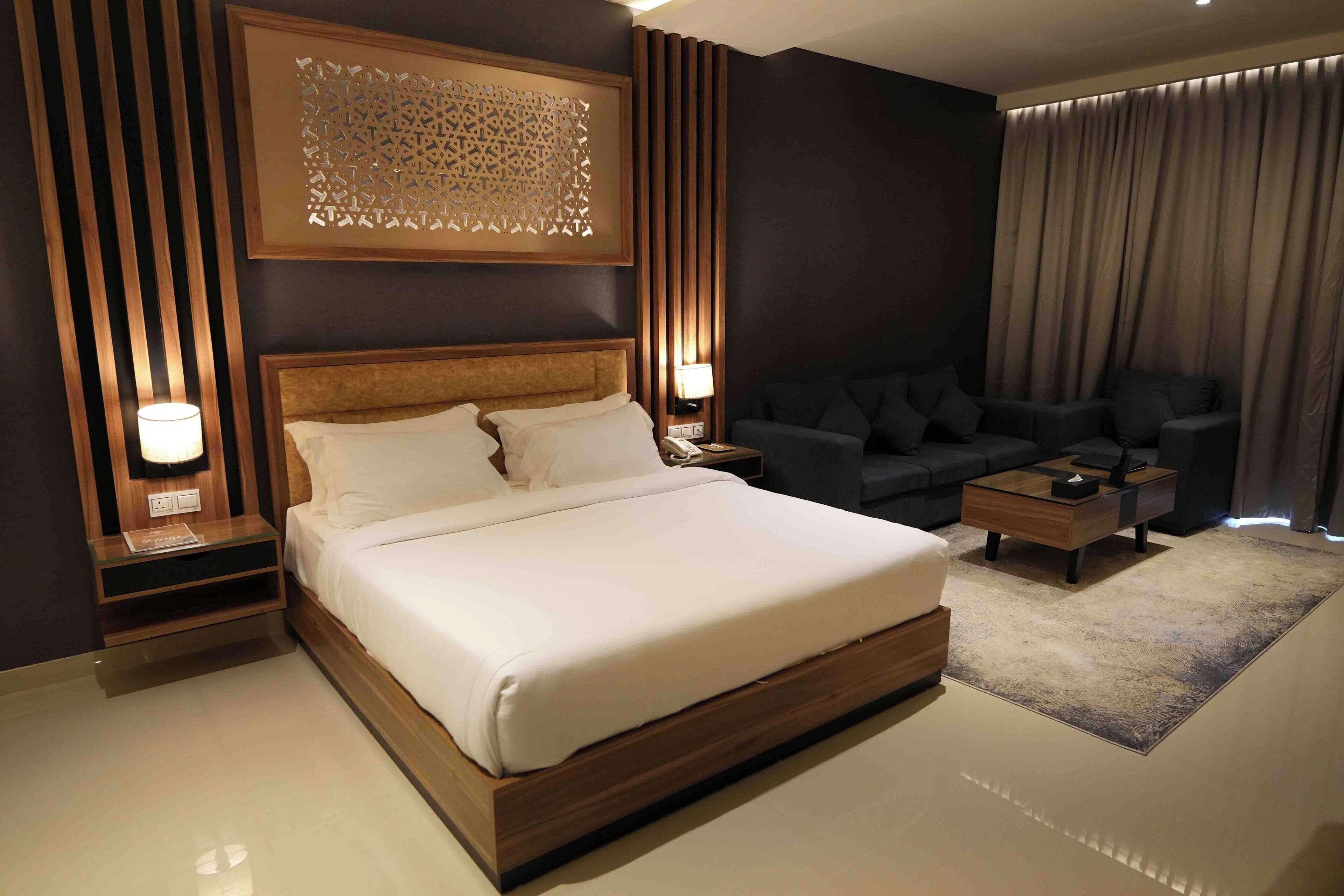 A modern hotel room with a large bed, a sofa, a coffee table, and a decorative wall panel.