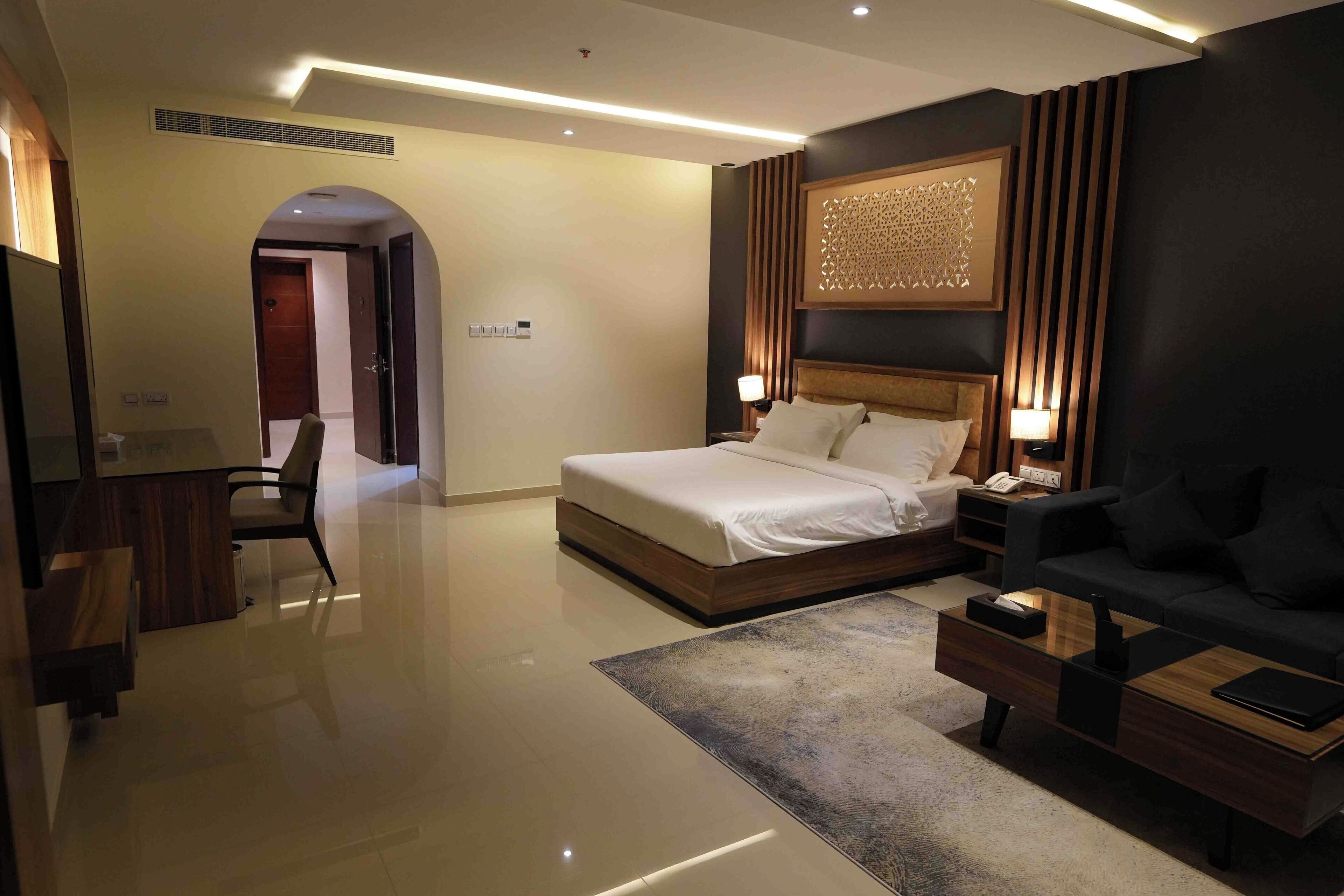 A modern hotel room with a large bed, a sitting area with a sofa, a desk, and a TV.