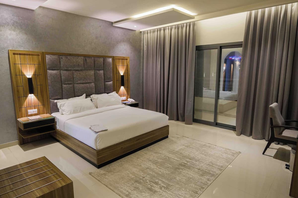 Una habitación de hotel moderna con una cama grande, cabecera de madera, lámparas de noche, una puerta de cristal que da a un balcón y un escritorio pequeño con una silla.