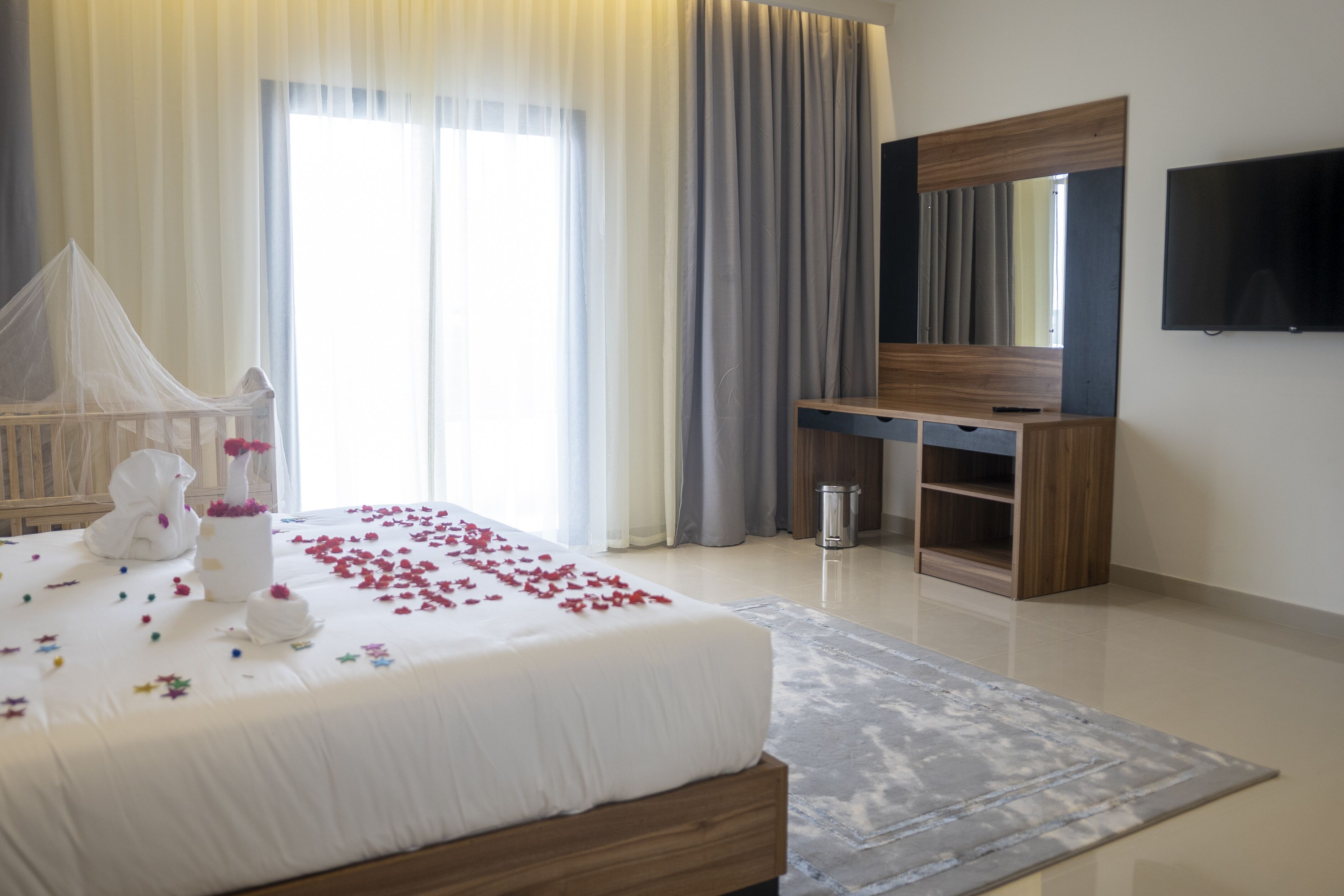 A hotel room with a bed, a TV, a desk, and a large window with curtains.