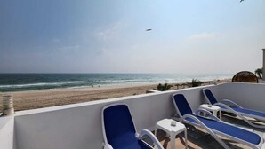 Un balcón con sillones reclinables azules y una pequeña mesa, con vista a una playa, el océano y un cielo despejado.