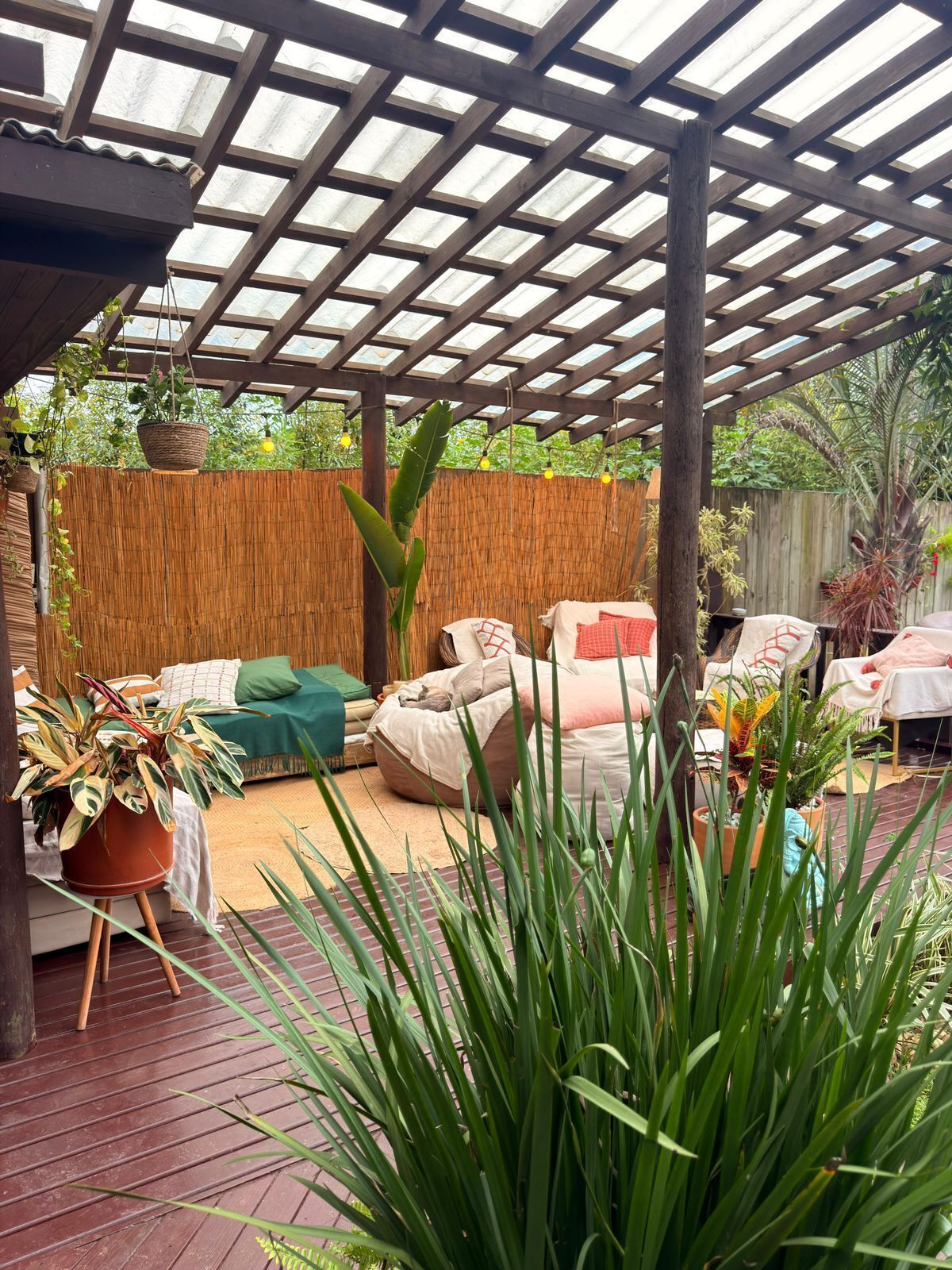 A covered patio area with a wooden pergola, outdoor seating with cushions, and various potted plants.
