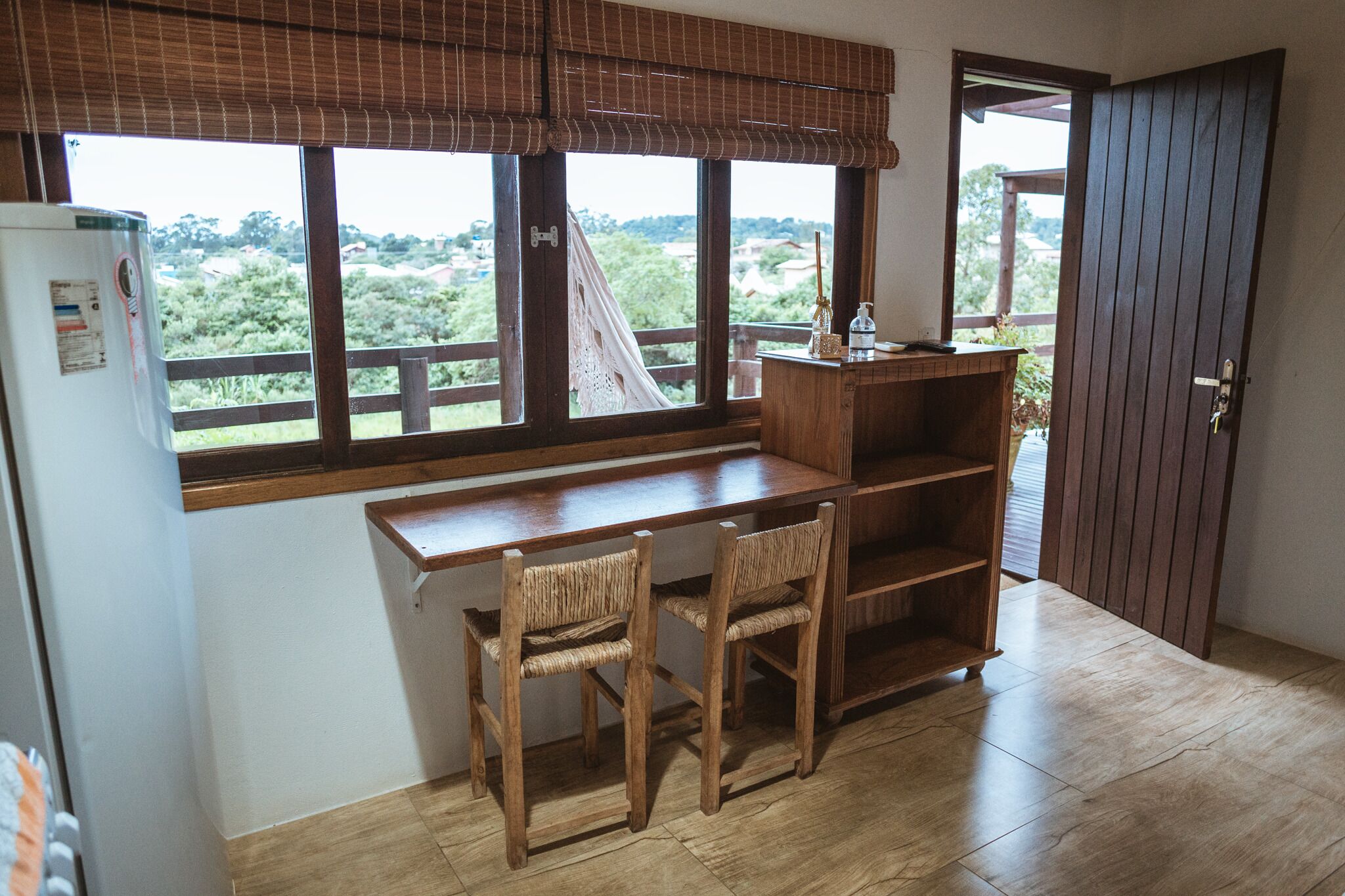 Quarto com escrivaninha de madeira, duas cadeiras de vime, uma estante de madeira e vista para o verde através de grandes janelas.