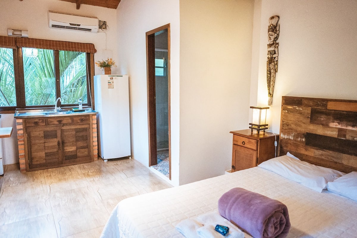Une pièce comprenant un lit, une table de chevet, une tête de lit en bois, une fenêtre donnant sur des palmiers, un coin cuisine avec un réfrigérateur et une porte menant à une autre pièce.