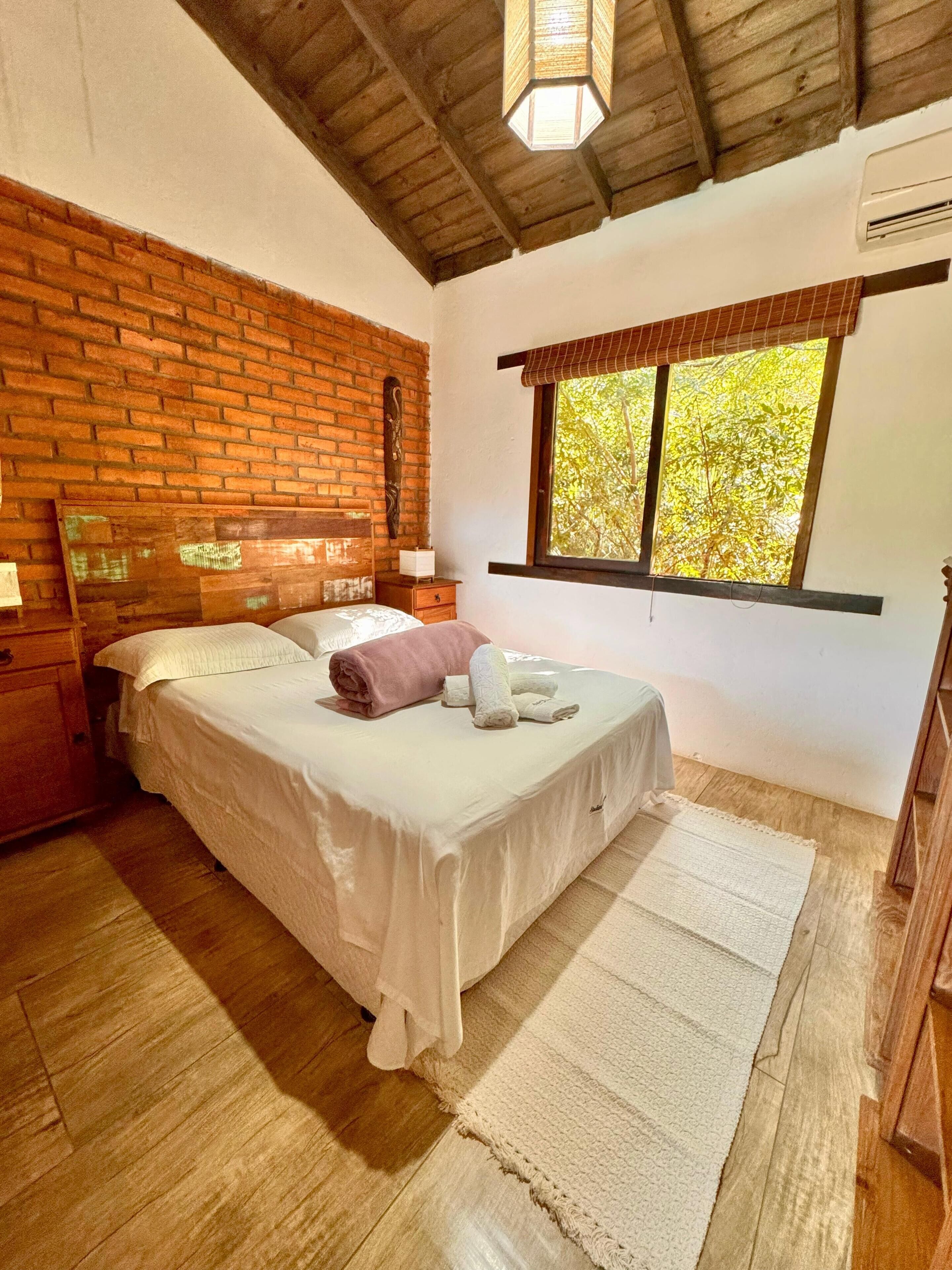 A bedroom with a brick wall, wooden ceiling, a bed with white bedding, a nightstand, a window with a view of trees, and a wooden floor.