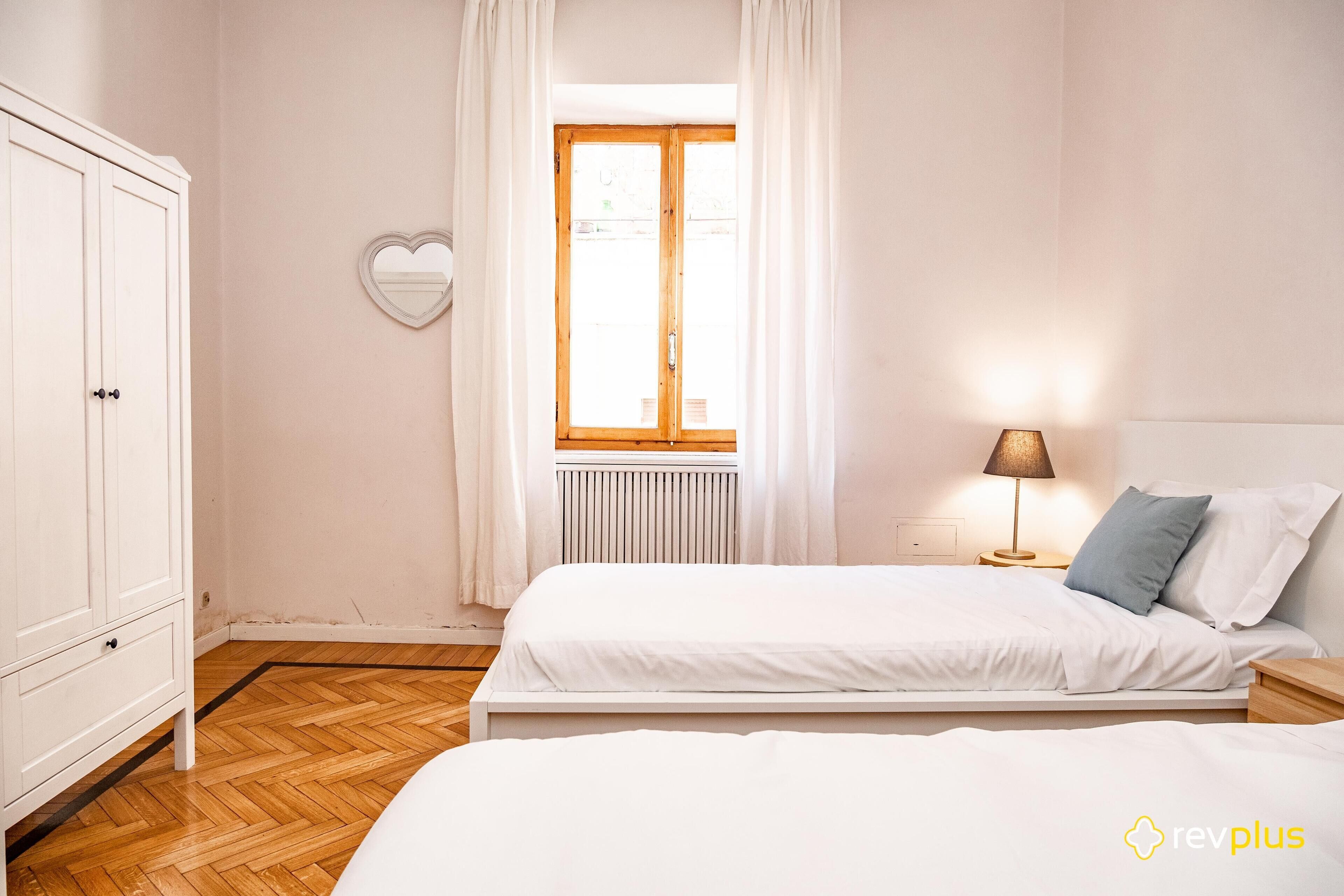 A bedroom with a bed, a wardrobe, a window with curtains, and a nightstand with a lamp.