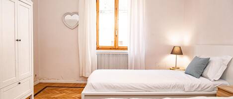 A bedroom with a bed, a wardrobe, a window with curtains, and a nightstand with a lamp.