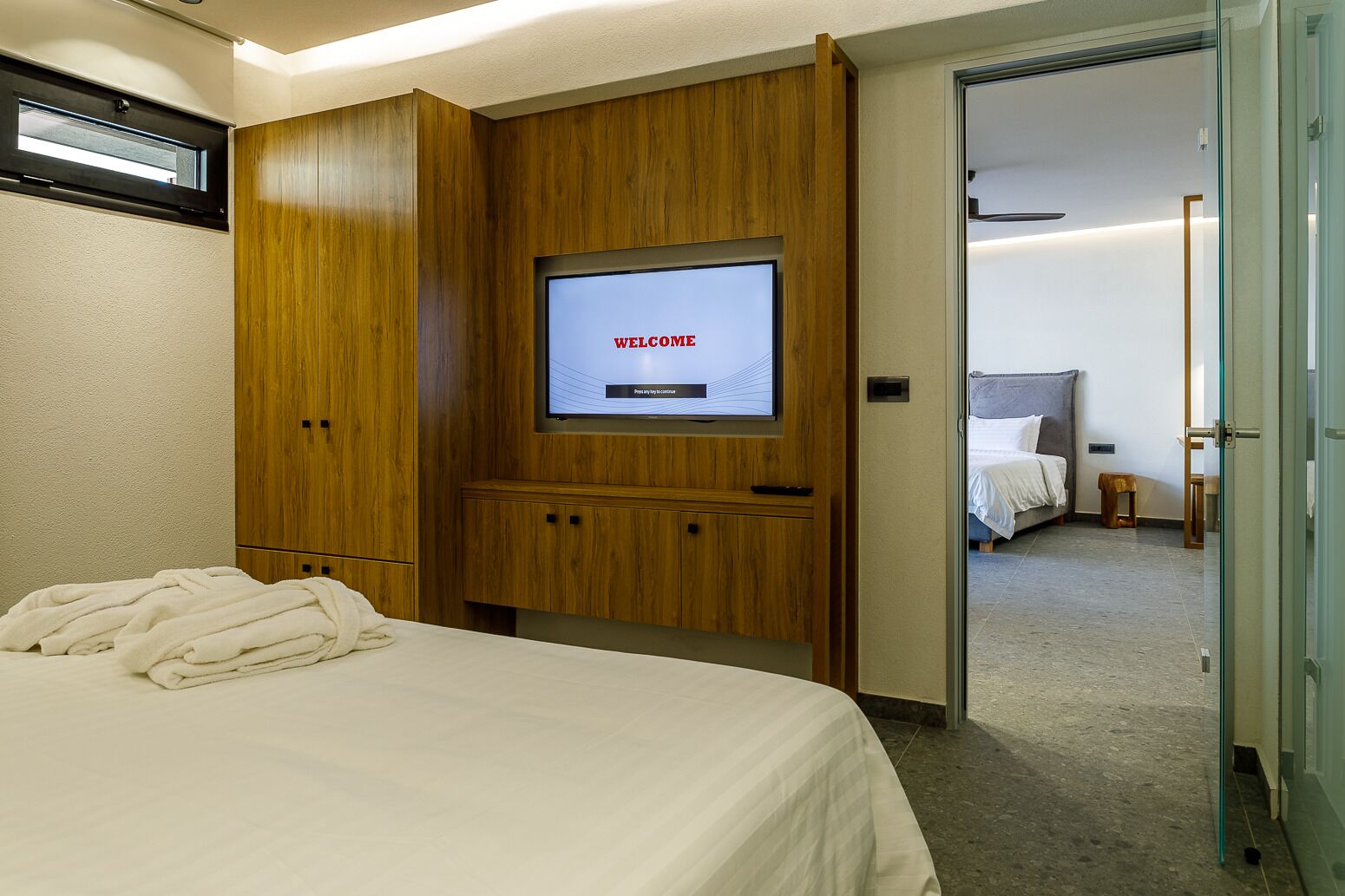 A hotel room with a bed, a TV screen displaying "WELCOME", wooden wardrobe, and a bathroom visible through an open door.