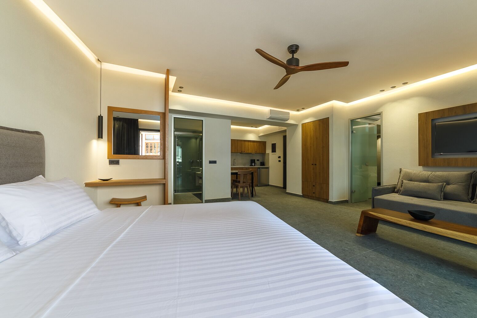 A modern hotel room with a large bed, a seating area with a sofa and a coffee table, a ceiling fan, and a kitchenette in the background.
