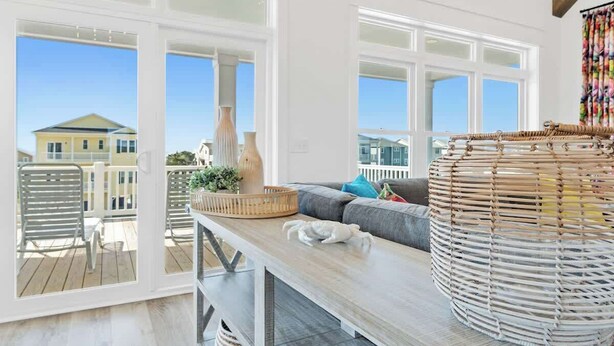A room with a large window, a wicker chair, a sofa with cushions, and a wooden table. - Big Orange Beach House by Brunswick Vacation Rentals: Heated Pool & Great Views (Ocean Isle Beach)
