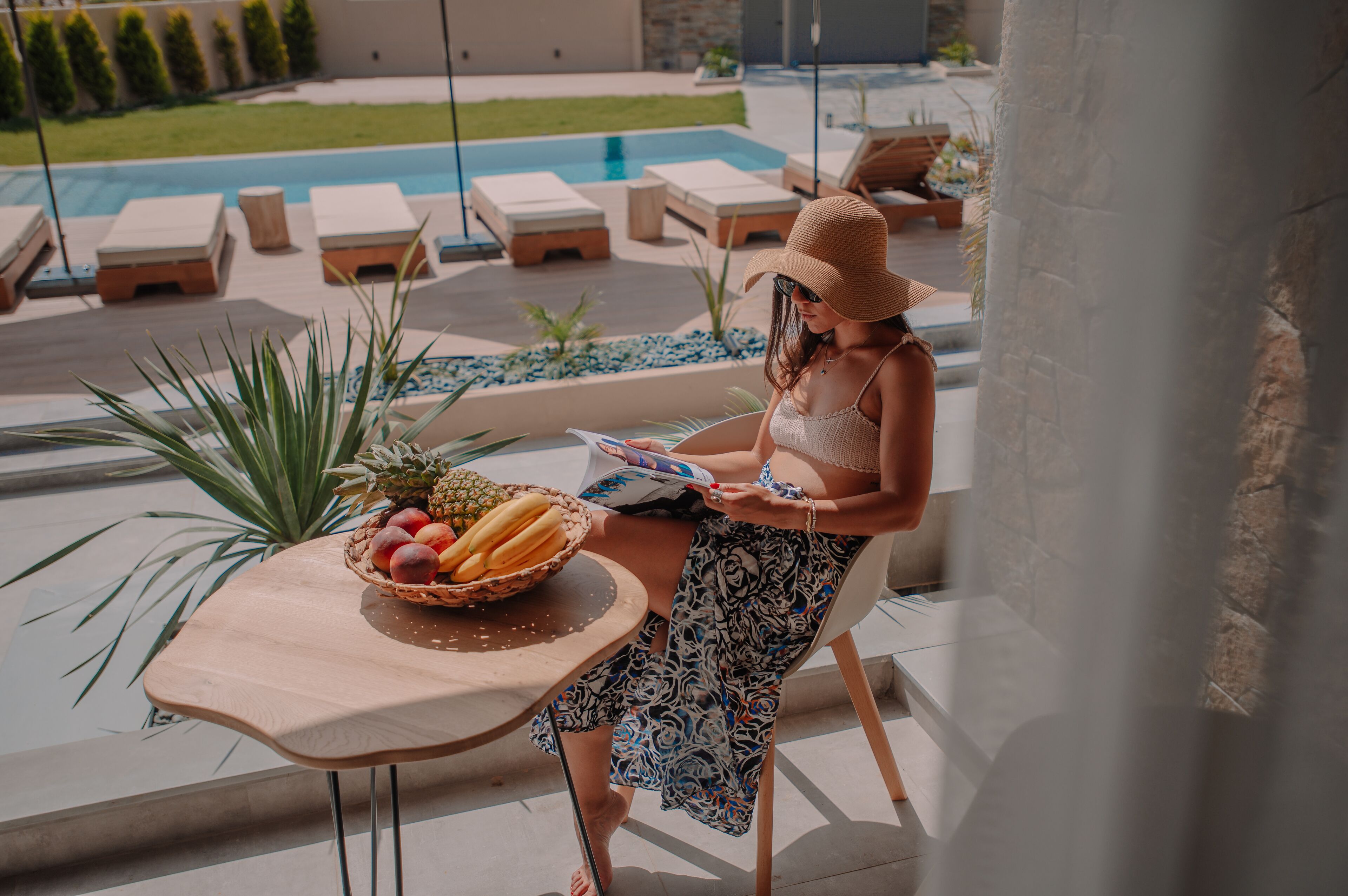Eine Person sitzt an einem Tisch mit einer Obstschale, blättert in einer Zeitschrift, in der Nähe eines Pools mit Liegestühlen und Blick auf einen Garten.