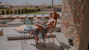 A woman relaxing by a poolside table with a drink, surrounded by lounge chairs and umbrellas. - Marble Luxury Suites (Heraklion)