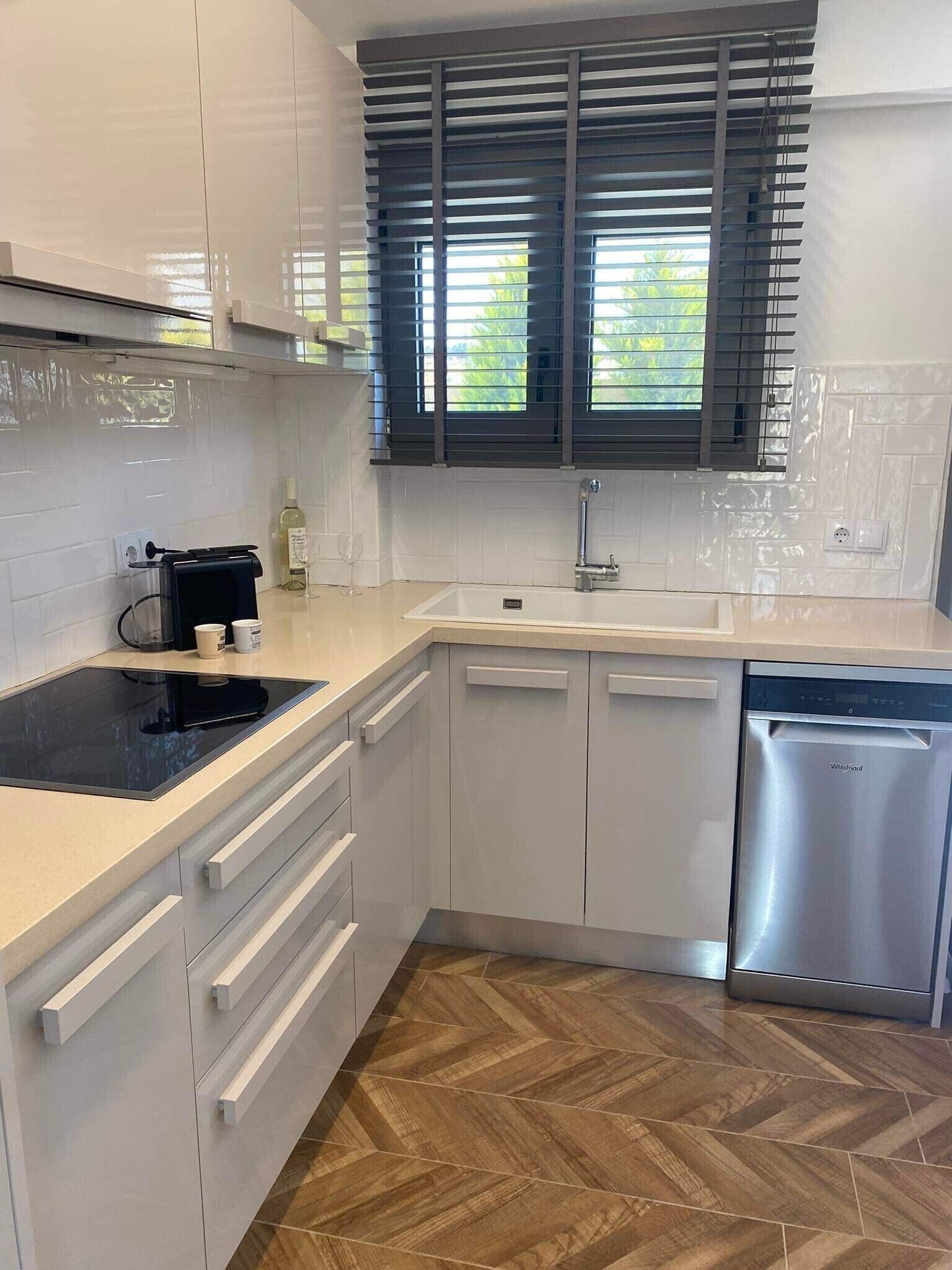 Une cuisine moderne avec des meubles blancs, une plaque de cuisson à induction noire, un lave-vaisselle en acier inoxydable et une fenêtre équipée de stores foncés.
