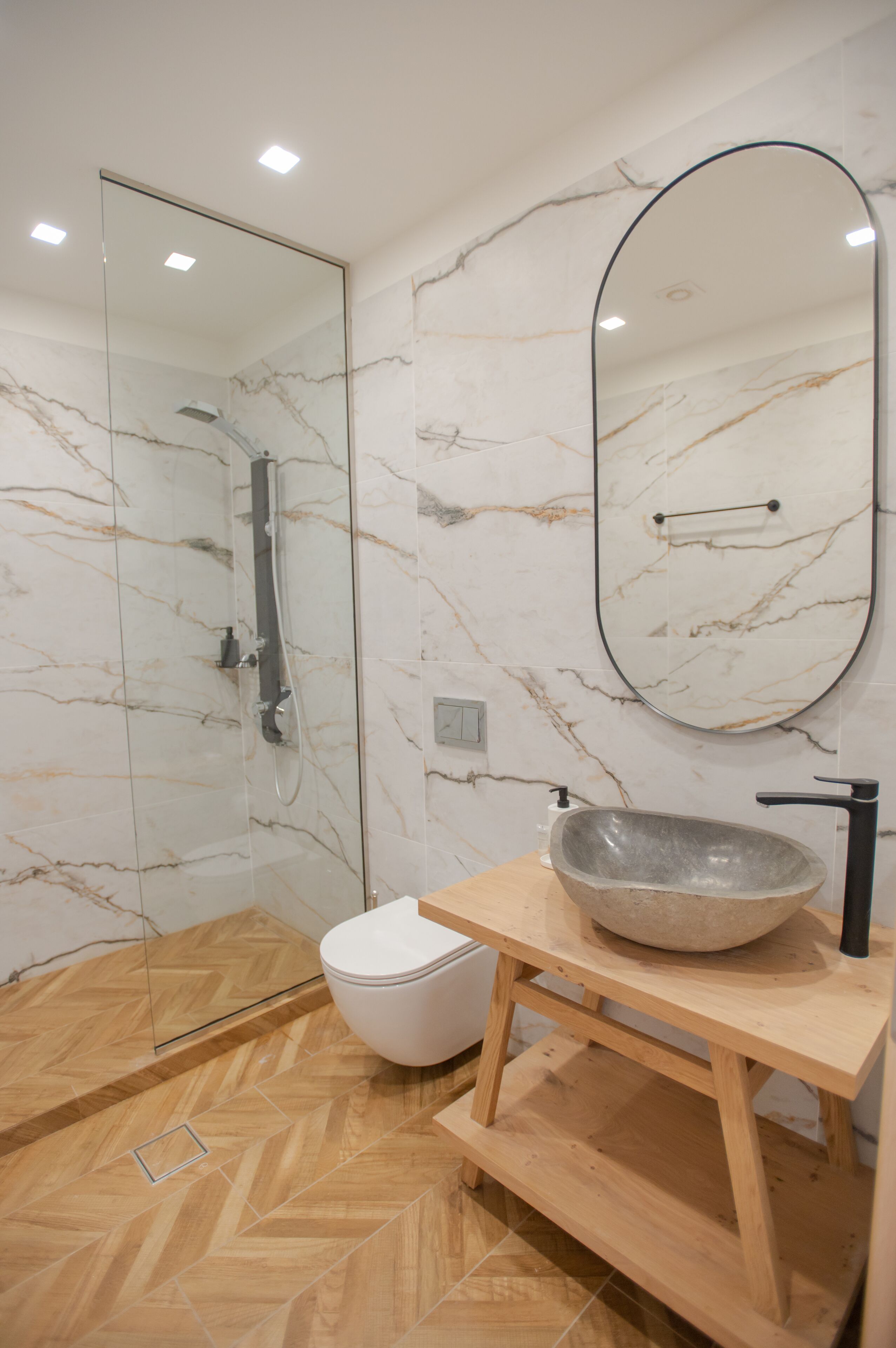Une salle de bain moderne avec une douche à parois de verre, un mur en marbre, un meuble-lavabo en bois et un grand miroir ovale.