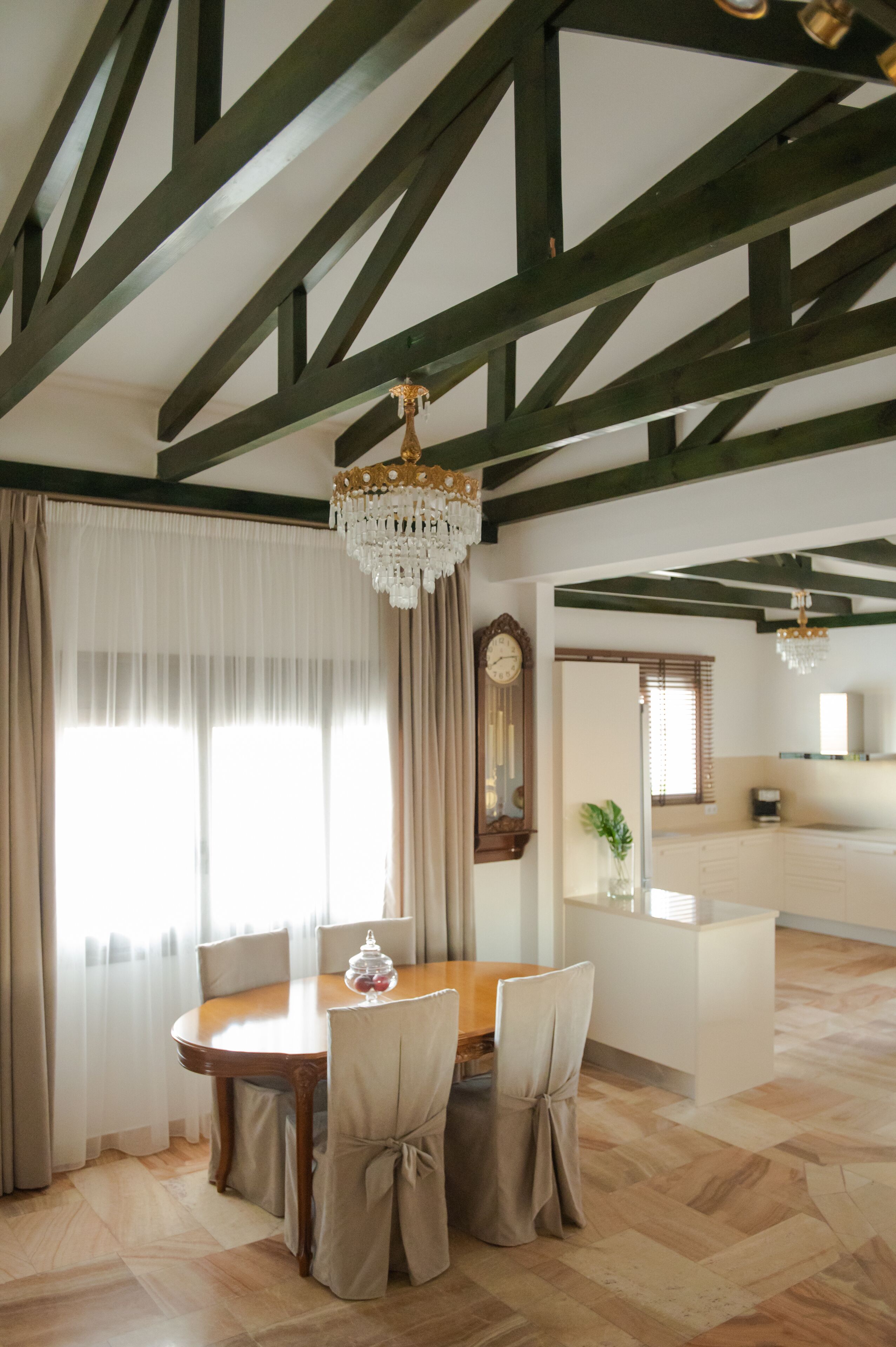 A dining area with a wooden table and chairs, a chandelier, and a large window with curtains.