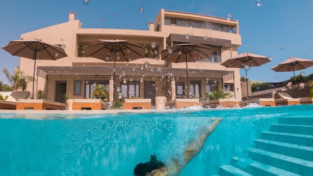 A swimming pool with a person diving, surrounded by umbrellas and a building with multiple floors and balconies. - Marble Luxury Suites (Heraklion)