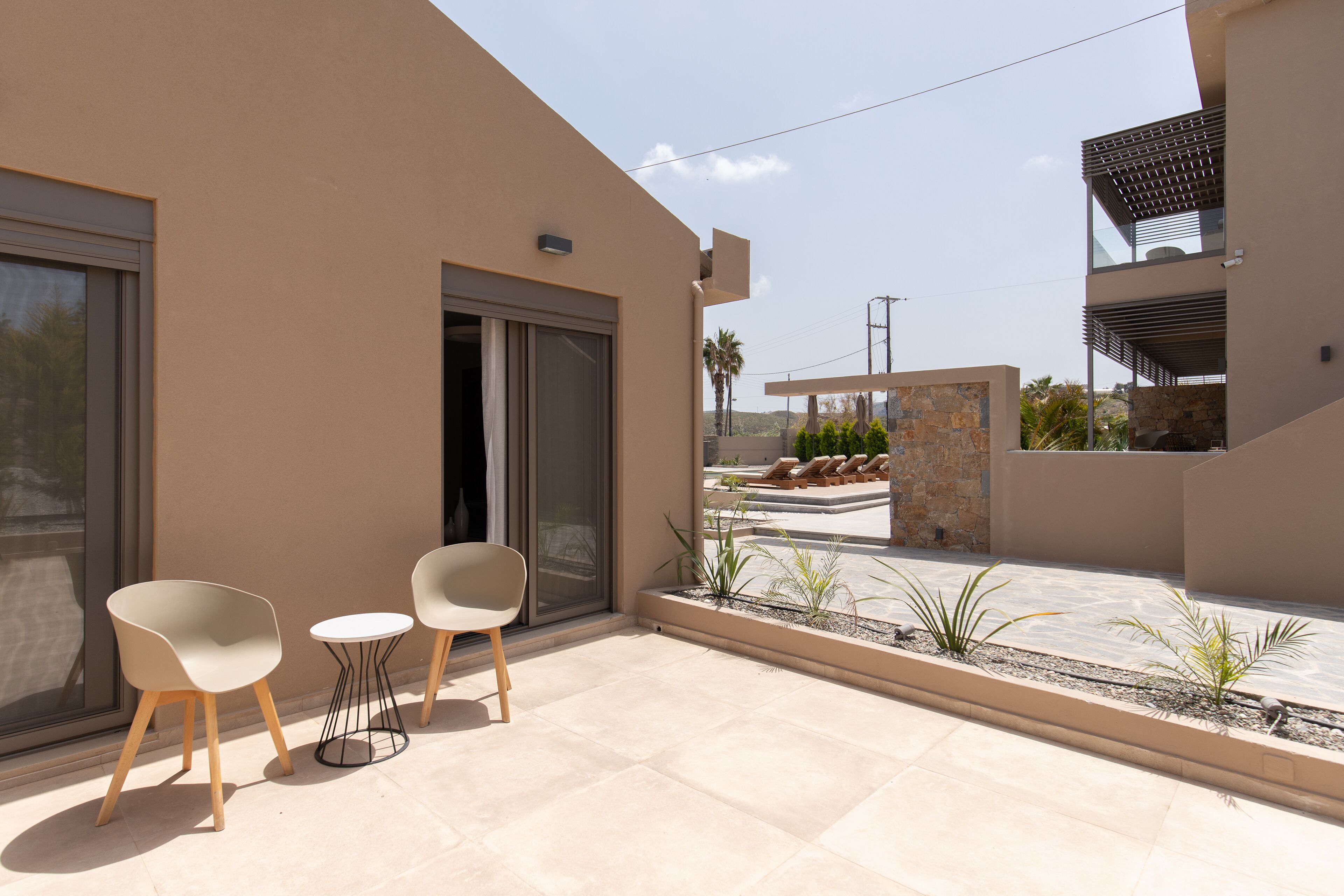 A modern outdoor patio with two chairs and a small table, a sliding glass door, and a view of a pool and palm trees.