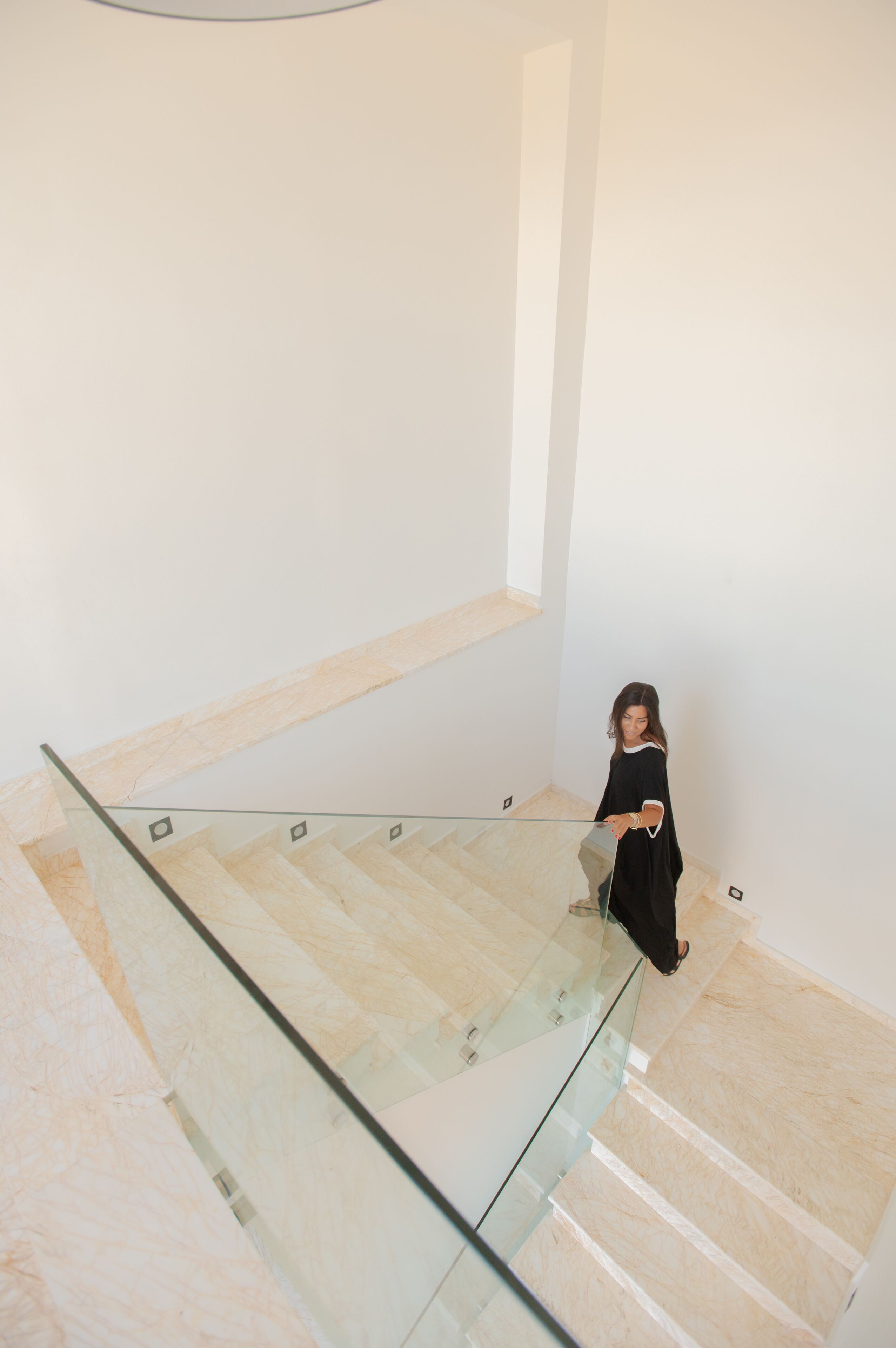 A person descending a staircase with a glass railing, in a modern interior with plain walls and a light-colored floor.