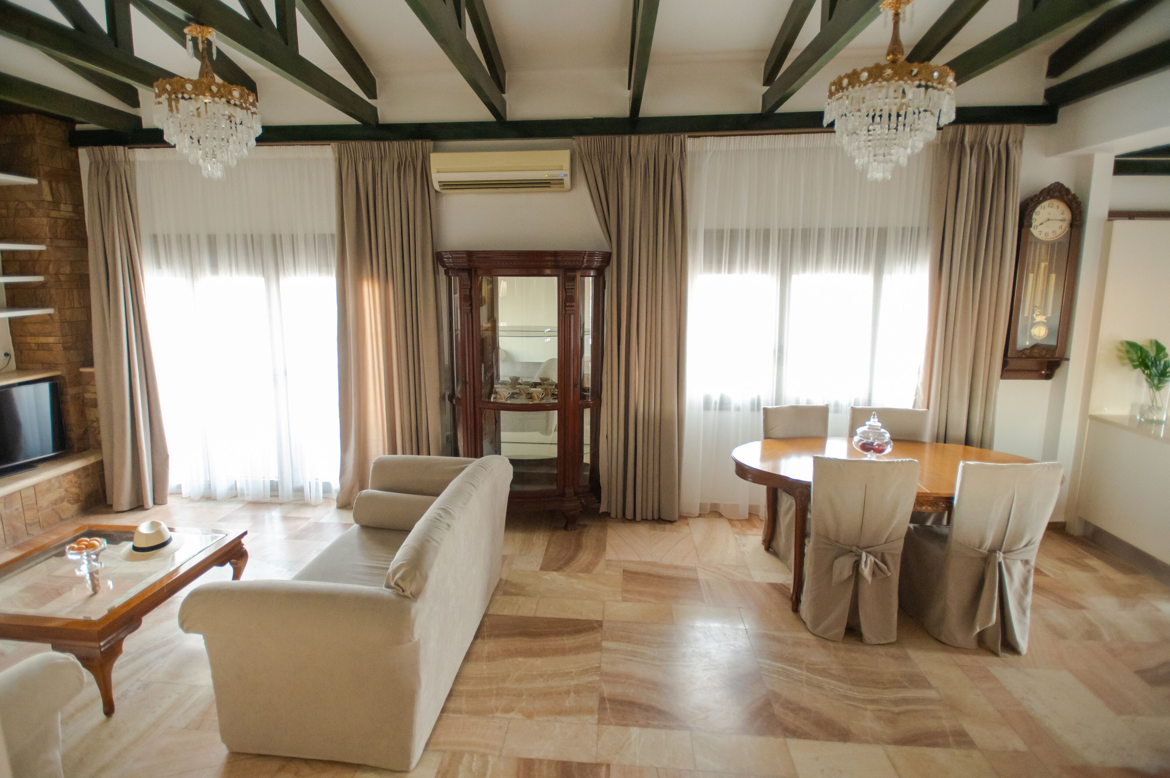 A spacious living room with a sofa, dining table, and chairs. There is a TV on the left and a chandelier hanging from the ceiling.
